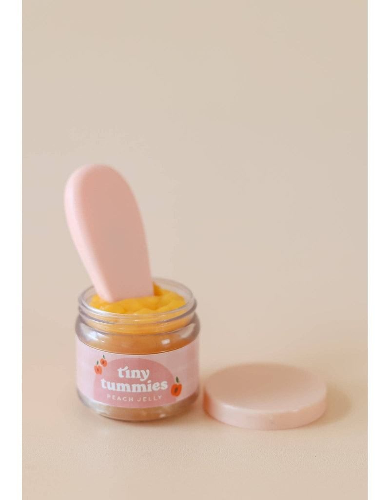 A small jar of "Tiny Tummies - Peach Jelly Food" from Tiny Harlow sits open, revealing the peach-colored jelly that's perfect for doll food. Inside is a magical spoon, and the lid rests nearby on a light neutral background.