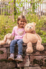 A smiling young girl sits on a stone wall amid greenery, holding a large teddy bear to her right and a smaller stuffed rabbit to her left. She wears jeans and a pink shirt with a teddy design, with a Steiff Berno Goldendoodle Dog Plush Stuffed Animal beside her.