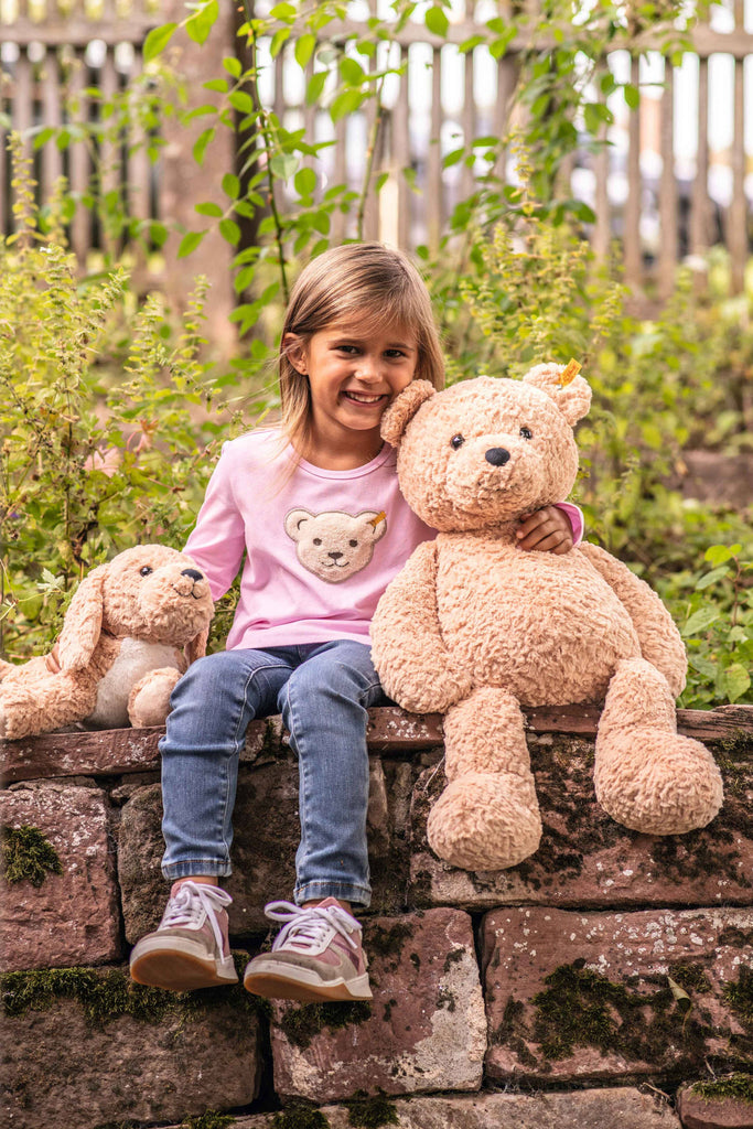 A smiling young girl sits on a stone wall amid greenery, holding a large teddy bear to her right and a smaller stuffed rabbit to her left. She wears jeans and a pink shirt with a teddy design, with a Steiff Berno Goldendoodle Dog Plush Stuffed Animal beside her.