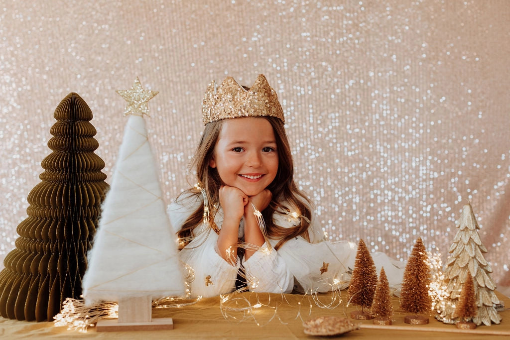 A young girl with long hair smiles at the camera, resting her chin on her hands. She wears a sparkly pink sequin crown and a white sweater. In front of her, there are various Christmas tree decorations in gold and white, alongside a pink sequin wreath. The background is a glittery, sparkling curtain.