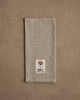 The Coral & Tusk Deck the Halls Bear Tea Towel rests on a textured brown surface, adorned with festive embroidery—ideal as a holiday kitchen towel for gatherings.
