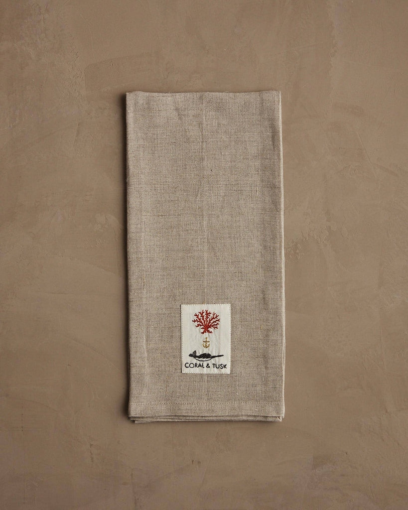The Coral & Tusk Deck the Halls Bear Tea Towel rests on a textured brown surface, adorned with festive embroidery—ideal as a holiday kitchen towel for gatherings.