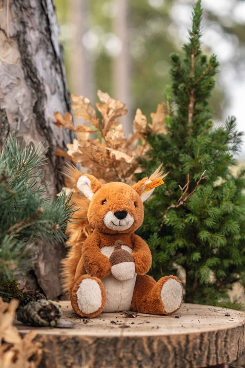 A Steiff, Phil Squirrel Stuffed Plush Animal, 8" with bushy tail sitting on a tree stump surrounded by greenery and autumn leaves in a forest setting, featuring a "Button in Ear.