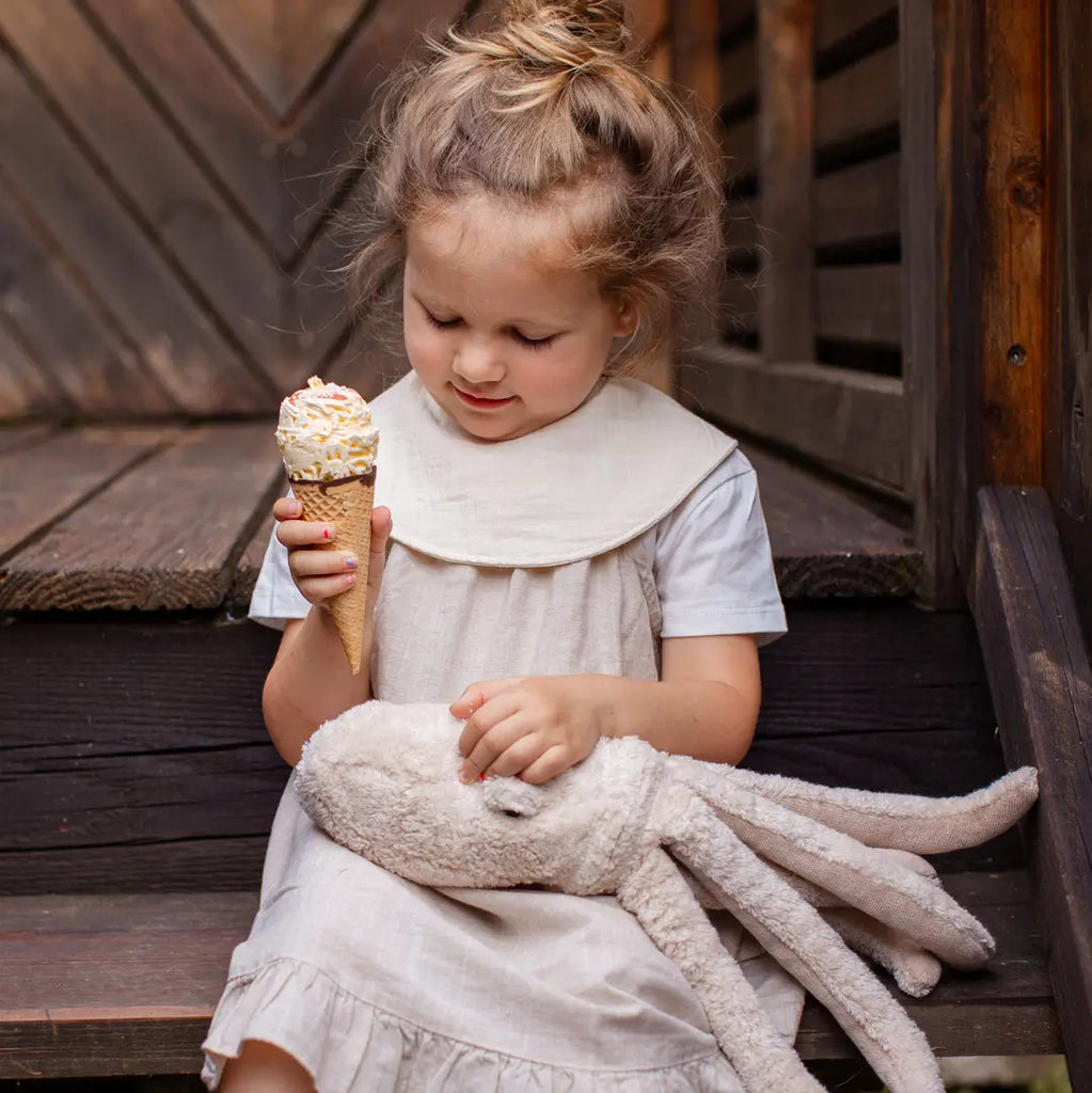 A young girl in a light dress sits on wooden steps, holding a Senger Naturwelt Cuddly Animal - Octopus for comfort as she admires an ice cream cone with sprinkles in her hand.