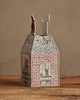 The Coral & Tusk Home for the Holidays Tissue Box Cover is a decorative, house-shaped cover with brick, window, and holiday light embroidery plus two festive figures on the roof, displayed on a wooden surface against a beige background.