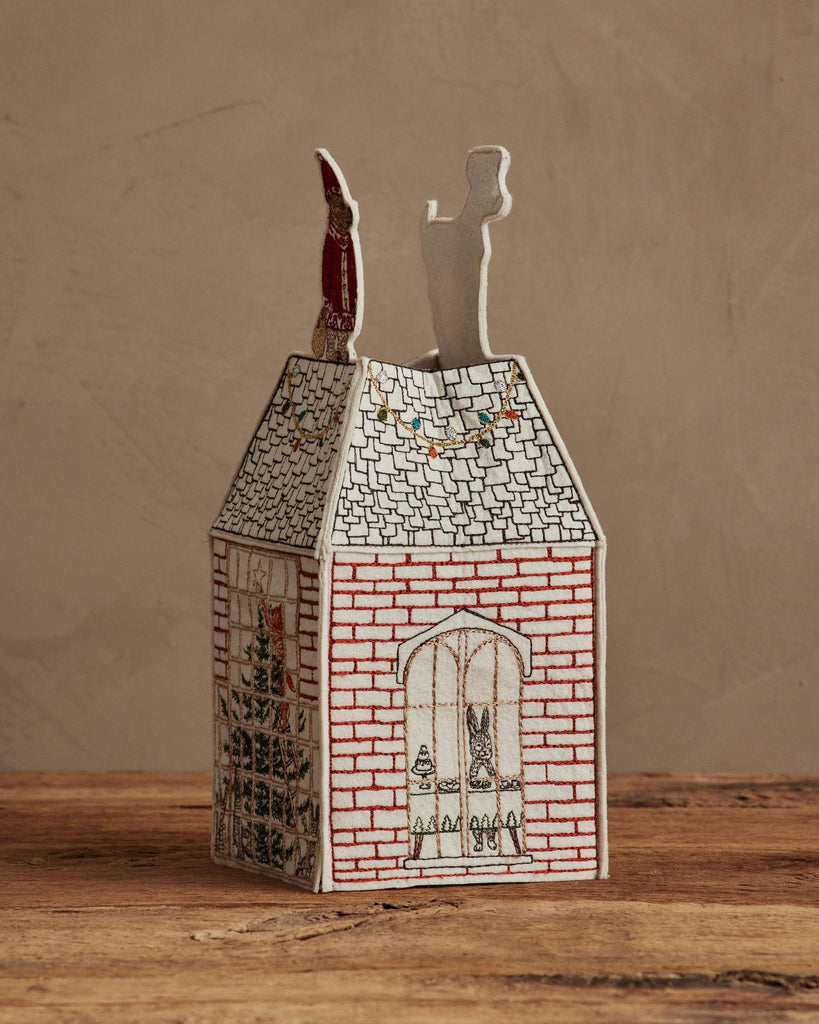 The Coral & Tusk Home for the Holidays Tissue Box Cover is a decorative, house-shaped cover with brick, window, and holiday light embroidery plus two festive figures on the roof, displayed on a wooden surface against a beige background.