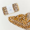 A folded and an opened Sarah's Silks Animal Print Playsilk - Cheetah, featuring yellow and black cheetah spots. The folded silk has a white label with blue text and a blue rainbow icon, ideal for Waldorf toys.