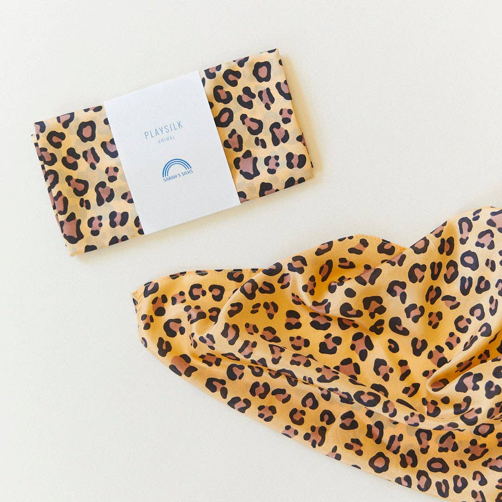 A folded and an opened Sarah's Silks Animal Print Playsilk - Cheetah, featuring yellow and black cheetah spots. The folded silk has a white label with blue text and a blue rainbow icon, ideal for Waldorf toys.