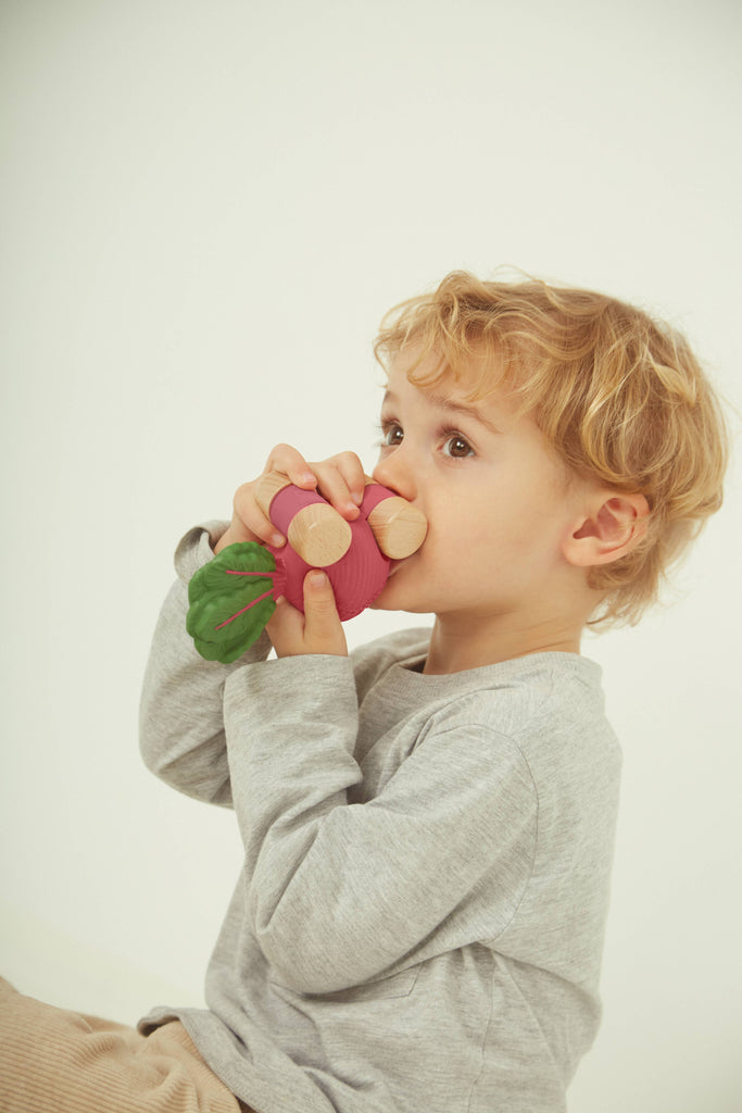 A young child with blond hair wearing a gray long-sleeve shirt holds and pretends to eat Betty The Beetroot Baby Car, a colorful FSC-certified wood toy.