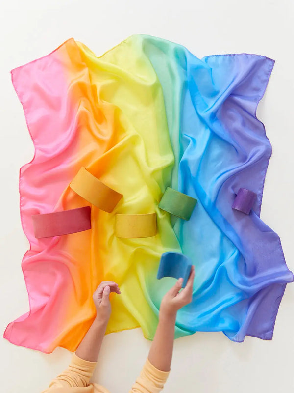 A child's hands are shown playing with a colorful, Sarah's Silk Enchanted Playsilk - Rainbow fabric that has several soft, velcro bands attached to it, displayed against a light background.