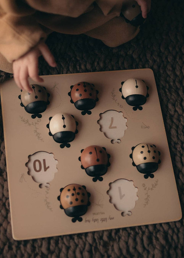 A child’s hand hovers over the My Little Lady Bug Wooden Puzzle—a Montessori counting toy featuring numbered slots and ladybug-shaped pieces in various shades of brown and beige. The board rests on a textured brown surface.