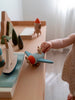 A small child in a polka dot outfit plays imaginatively with the Sabo Concept Aviator Mouse, a blue wooden airplane, sailboat, and trees on a light wooden table by the window.