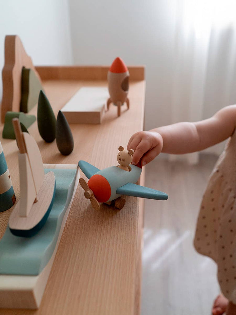 A small child in a polka dot outfit plays imaginatively with the Sabo Concept Aviator Mouse, a blue wooden airplane, sailboat, and trees on a light wooden table by the window.