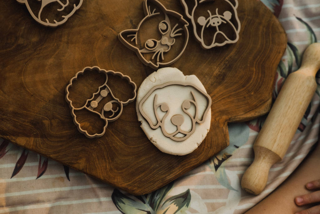 The Pet Friends Faces Cutter Set – Eco Playdough Tool, featuring dog, cat, and bear-shaped cutters, rests on a wooden board with dough. One cutter presses into the dough to create a playful dog face—perfect for creative sensory play. A rolling pin lies close by on a patterned cloth.