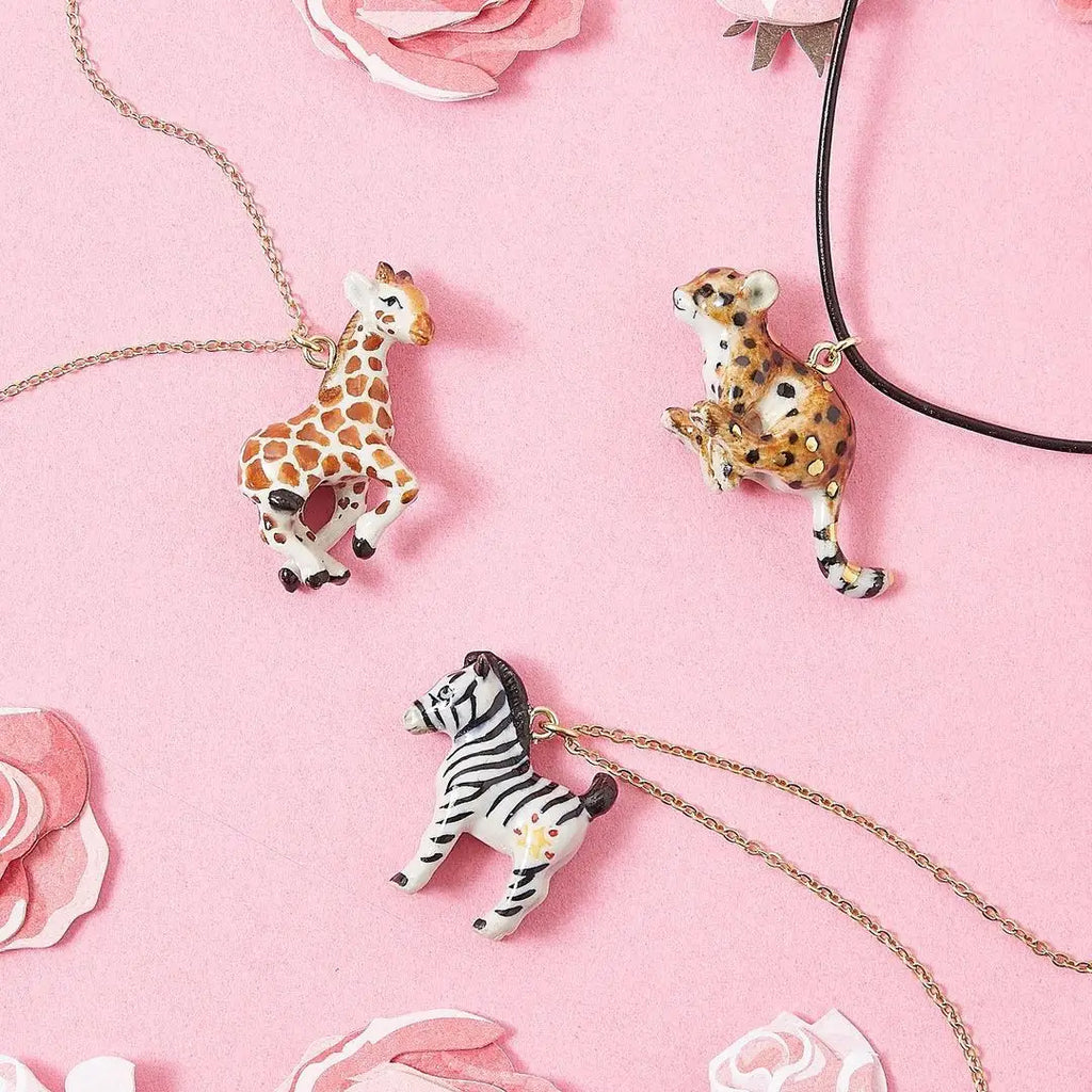 A photo of three hand-painted, animal-shaped necklaces on a pink background with decorative paper flowers. The pendants include a giraffe on a gold-plated chain, a cheetah on a black leather cord, and a Zebra Necklace.