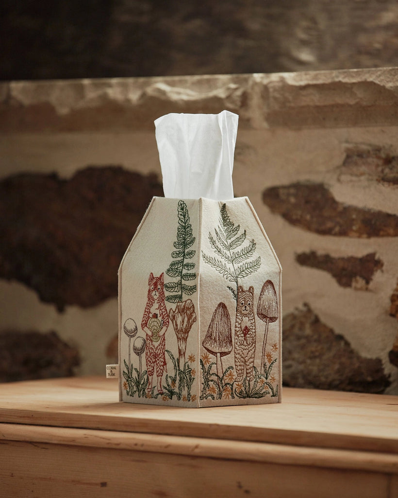 A Coral & Tusk Mushrooms and Ferns tissue box cover designed to resemble a house, featuring intricate embroidery of woodland elements like a fox, ferns, and mushrooms on a cream fabric, placed on a wooden surface.