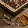 A Scrabble Heirloom with Rotating Game Board sits on the table, with letter tiles on racks spelling “SCORE BG” and “WINNER,” while a brown pouch nearby spills scattered tiles.