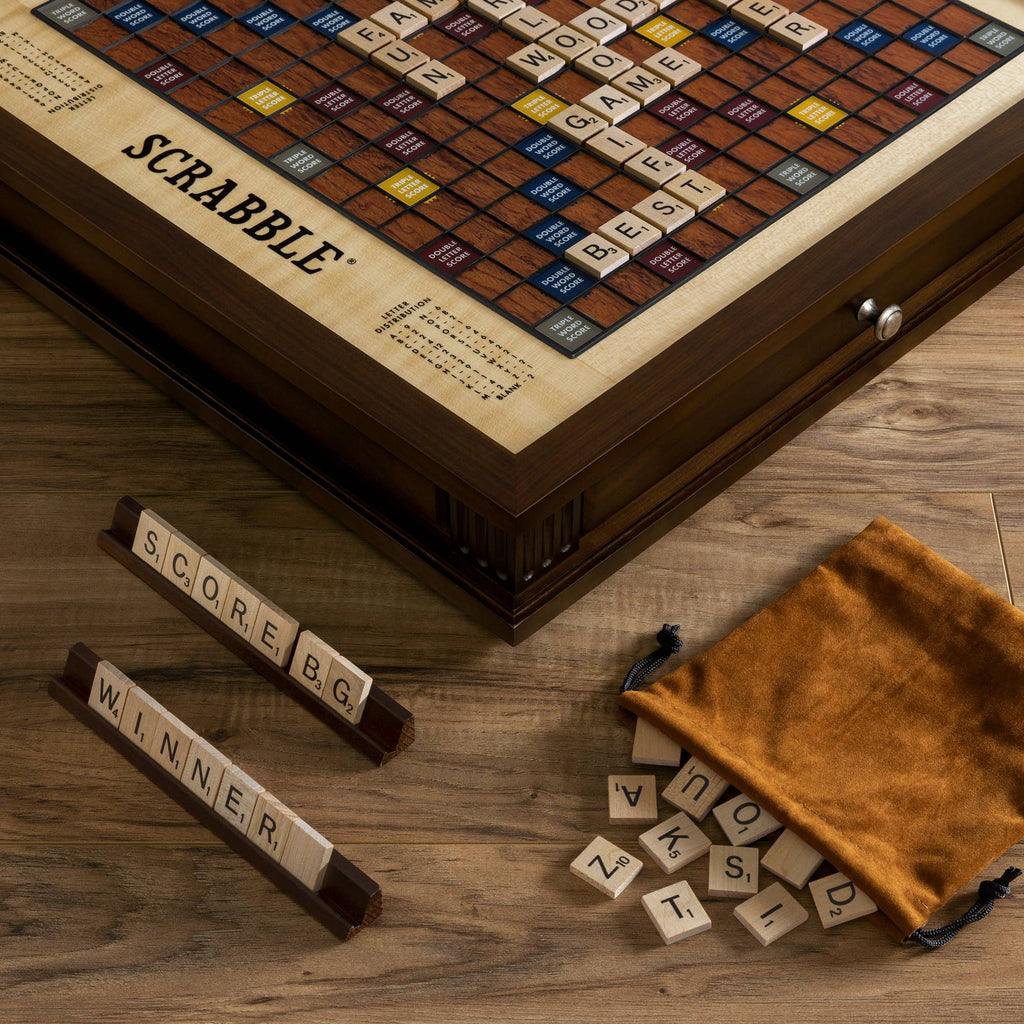 A Scrabble Heirloom with Rotating Game Board sits on the table, with letter tiles on racks spelling “SCORE BG” and “WINNER,” while a brown pouch nearby spills scattered tiles.