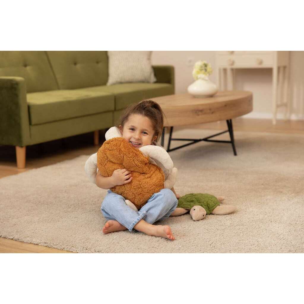A young girl sits barefoot on a beige carpet, smiling and hugging a plush toy with the Senger Naturwelt Cuddly Animal - Turtle beside her. The background features a green sofa and wooden table.
