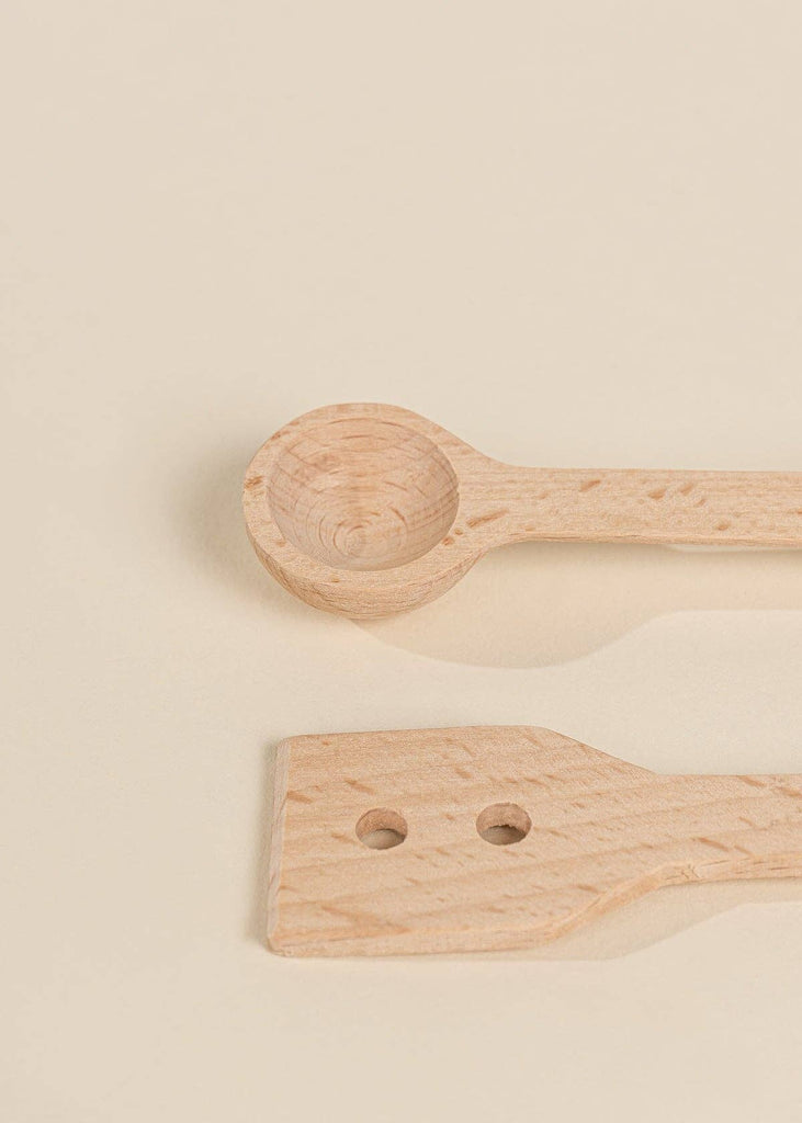 Two wooden utensils from the Wooden Pots and Pans Playset (6 Pcs) rest on a light beige surface: one features a round, spoon-like end, while the other has a flat, angled end with two small holes—ideal additions to any kids kitchen playset or collection of wooden cooking toys.