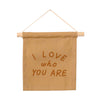 The I Love Who You Are Canvas Sign - Peach features "I LOVE who YOU ARE" in bold brown on natural tan canvas, handmade by Kenyan artisans and hung from a wooden dowel with string—perfect for heartfelt display.