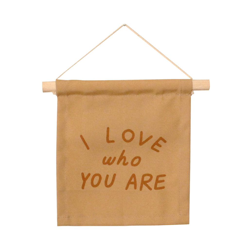 The I Love Who You Are Canvas Sign - Peach features "I LOVE who YOU ARE" in bold brown on natural tan canvas, handmade by Kenyan artisans and hung from a wooden dowel with string—perfect for heartfelt display.