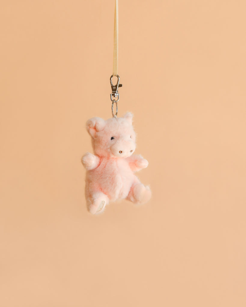 The Bukowski - Baby Oink Pig Bag Charm, a small plush pink piglet keychain, hangs from a beige strap and metal clasp against a plain peach-colored background, making it the perfect plush accessory for your bag.