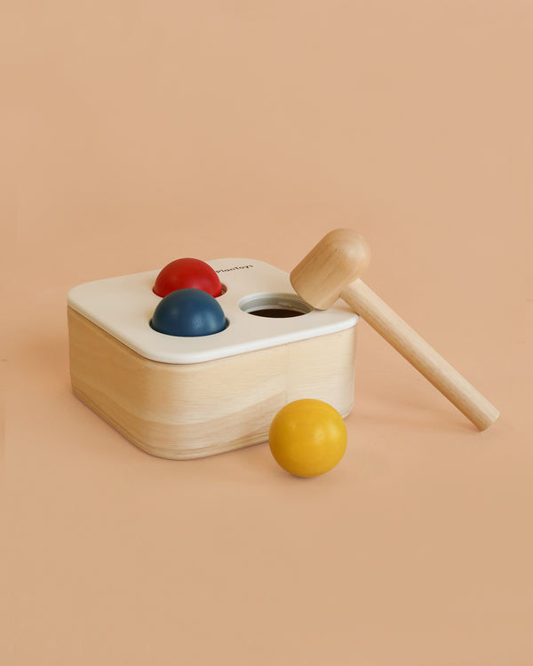 The PlanToys Punch & Drop Bin is a wooden cause and effect toy featuring three colorful balls in red, blue, and yellow, a top hole for dropping balls, and a wooden mallet—displayed against a peach background.