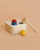 The PlanToys Punch & Drop Bin is a wooden cause and effect toy featuring three colorful balls in red, blue, and yellow, a top hole for dropping balls, and a wooden mallet—displayed against a peach background.