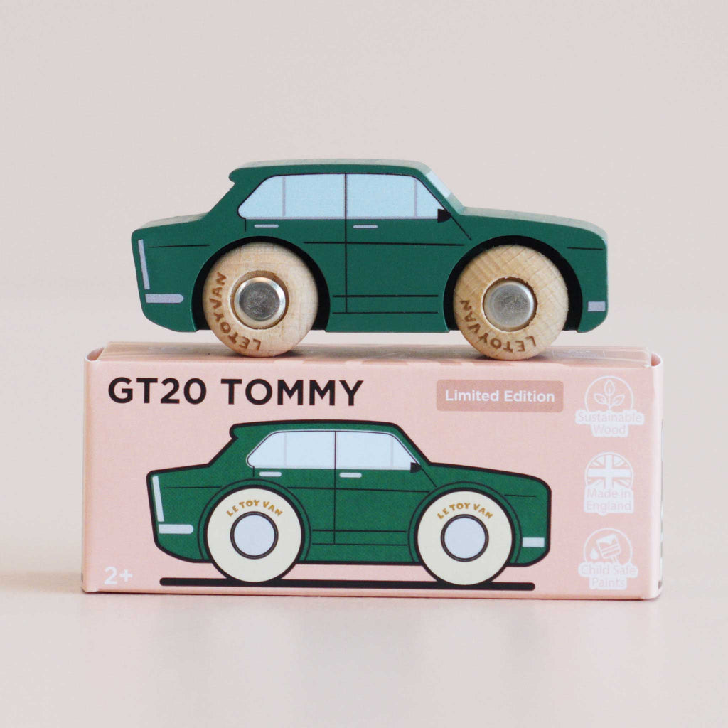 Revamped sentence: A nostalgic toy, the green wooden car—No. 3 GT20 Tommy | Made in England—with “Le Toy Van” on its wheels, sits atop a pink box labeled “GT20 TOMMY,” featuring icons for sustainability, British design, and safety.