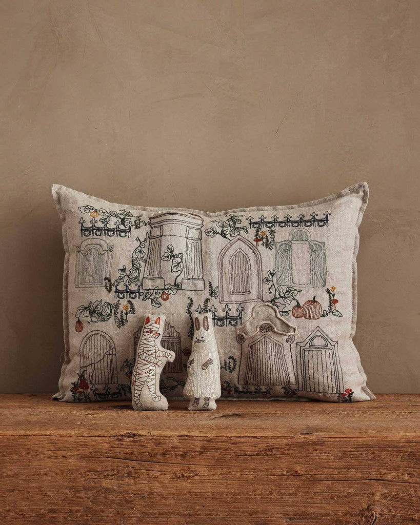 The Coral & Tusk Graveyard Pocket Pillow, featuring embroidered doors and plants, leans against a beige wall with two small fabric animal dolls, including a Bunny Ghost, displayed in front.