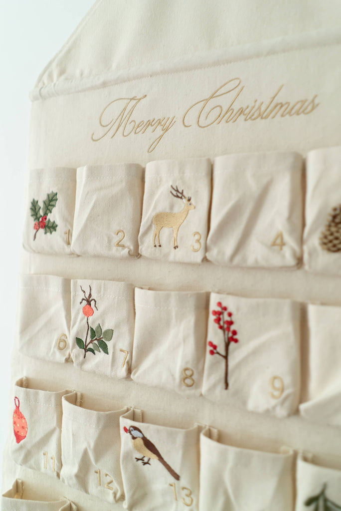 The Sabo Concept Christmas Cotton Advent Calendar features "Merry Christmas" embroidery and 12 numbered pockets, each decorated with festive designs such as a deer, pinecone, holly, berries, ornament, and bird.