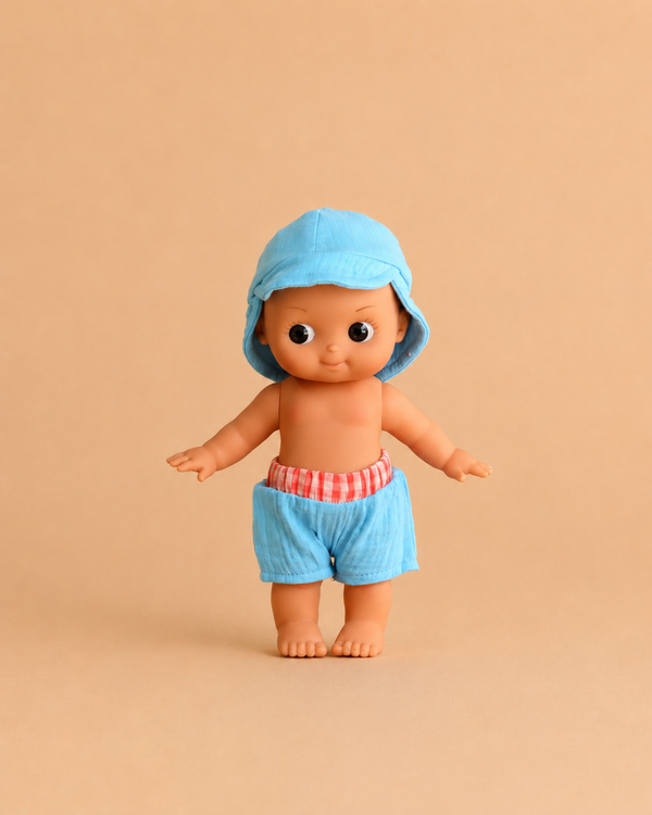 The Minikane Mini Pancha Doll - Basile with Riviera Blue Outfit, featuring big eyes, a blue hood, and blue shorts with a red-and-white waistband, stands against a plain beige background—perfect for imaginative play and crafted from high-quality vinyl.
