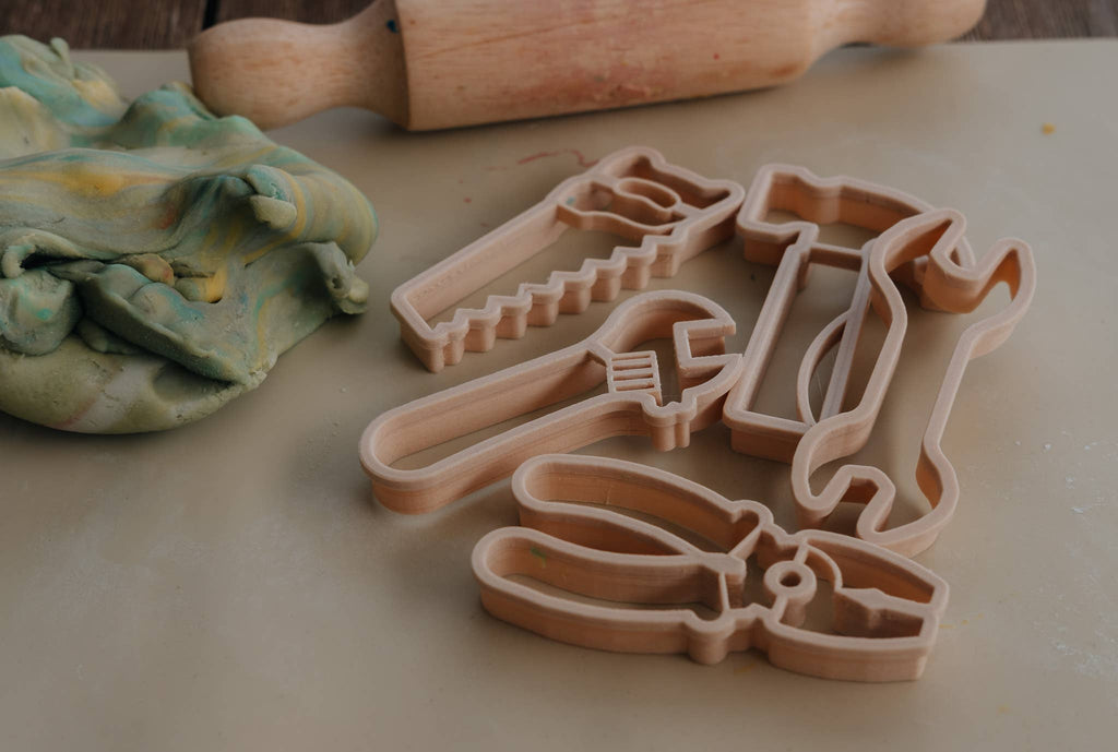The Tools Cutter Set - Eco Playdough Tool features wooden cookie cutters shaped like a saw, hammer, wrench, pliers, and spanner, shown here beside a rolling pin and some greenish dough on a beige surface. This eco-friendly tool-themed set is perfect for creative baking and sustainable play.