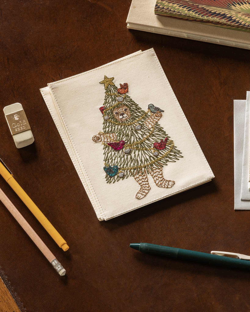 A Coral & Tusk Christmas Tree Bear Card features an embroidered bear dressed in a Christmas tree costume with ornaments and birds, lying on a wooden desk with pencils and an eraser.