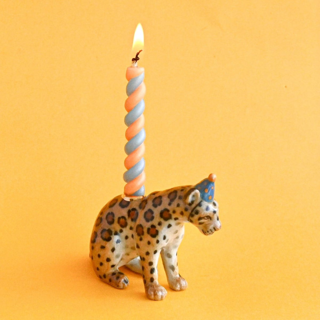 A small figurine of a jaguar, crafted from fine porcelain, wears a blue party hat with a lighted orange and blue striped candle inserted vertically into its back. Set against a solid orange background, this collectible Jaguar Cake Topper exudes heirloom quality charm.