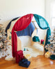 A cozy indoor play fort featuring Sarah's Silk Giant Evergreen Playsilk with red and teal drapes, surrounded by snow-dusted Christmas trees, has wrapped presents in vibrant winter colorways scattered around on a fluffy white rug and hardwood floor.