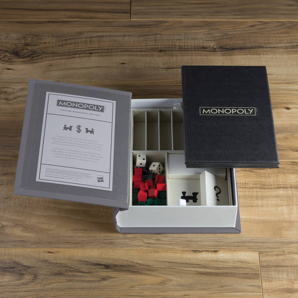 An open Monopoly Vintage Bookshelf Edition box sits on a wooden surface, displaying premium dice, cards, pieces, and instructions—all organized neatly inside its elegant book-shaped case.
