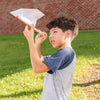 A boy stands on grass near a brick wall, holding the Huckleberry Paper Plane Launcher and aiming it upward, preparing to launch a paper airplane into the sky.