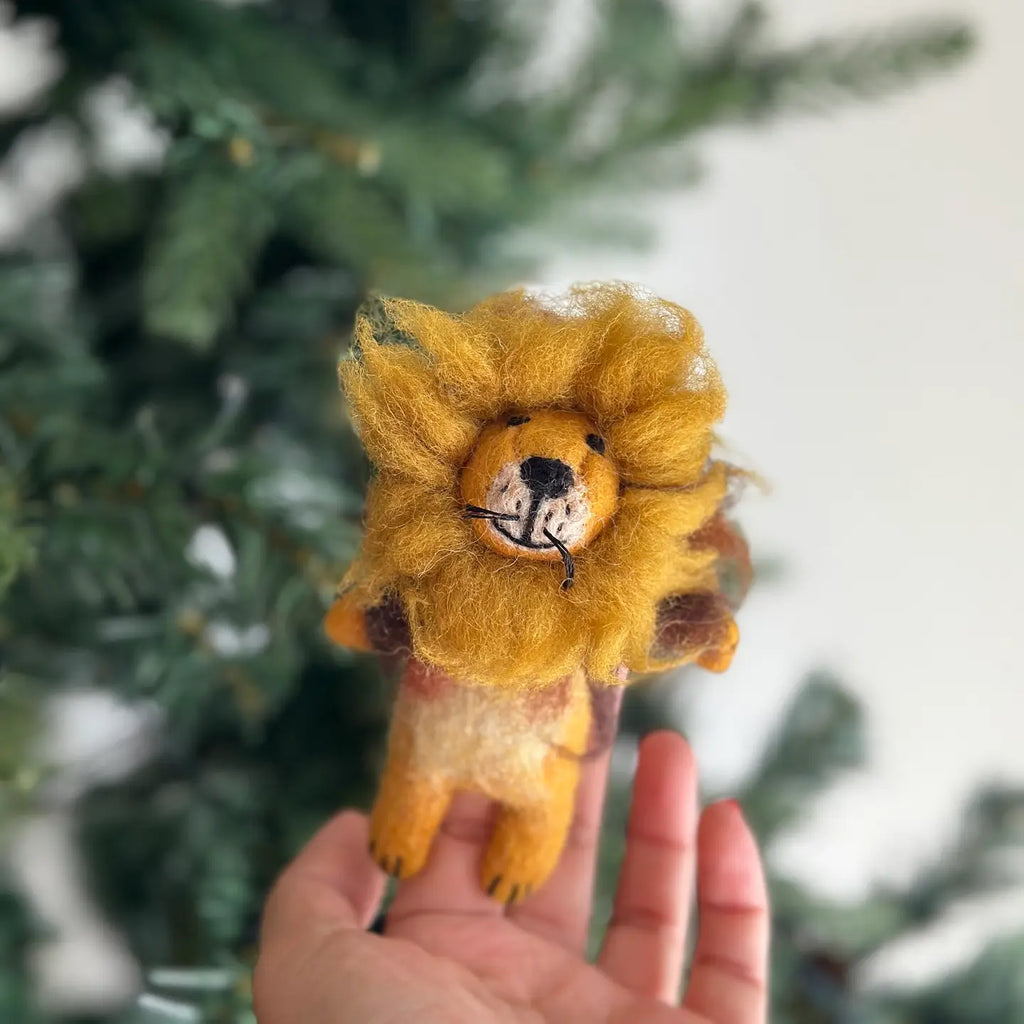 A hand holding the "Fluffy Lion Finger Puppet," a small felt lion with a yellow mane and an adorable facial expression. Handmade in Nepal, this charming figurine makes for an excellent storytelling prop. The background features some out-of-focus greenery, possibly a plant or tree, giving the image a natural look.