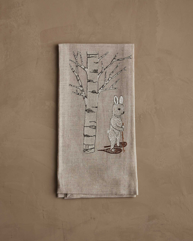 The Coral & Tusk Snowshoe Hare Tea Towel features a beige linen fabric with an embroidered bare aspen tree and a rabbit, set against a neutral brown textured background for a quiet winter charm.