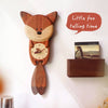 A Handmade Wooden Fox Wall Clock hangs beside a small shelf with a photo frame. The fox’s tail swings as the clock’s pendulum—ideal for nursery decor. Text reads: “Little fox telling time.”.