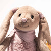 The Steiff Cowgirl Hoppie Rabbit is a soft, plush bunny with light brown fur, long floppy ears, black button eyes, a pink nose, a light pink scarf around its neck, and a yellow tag on its left ear.