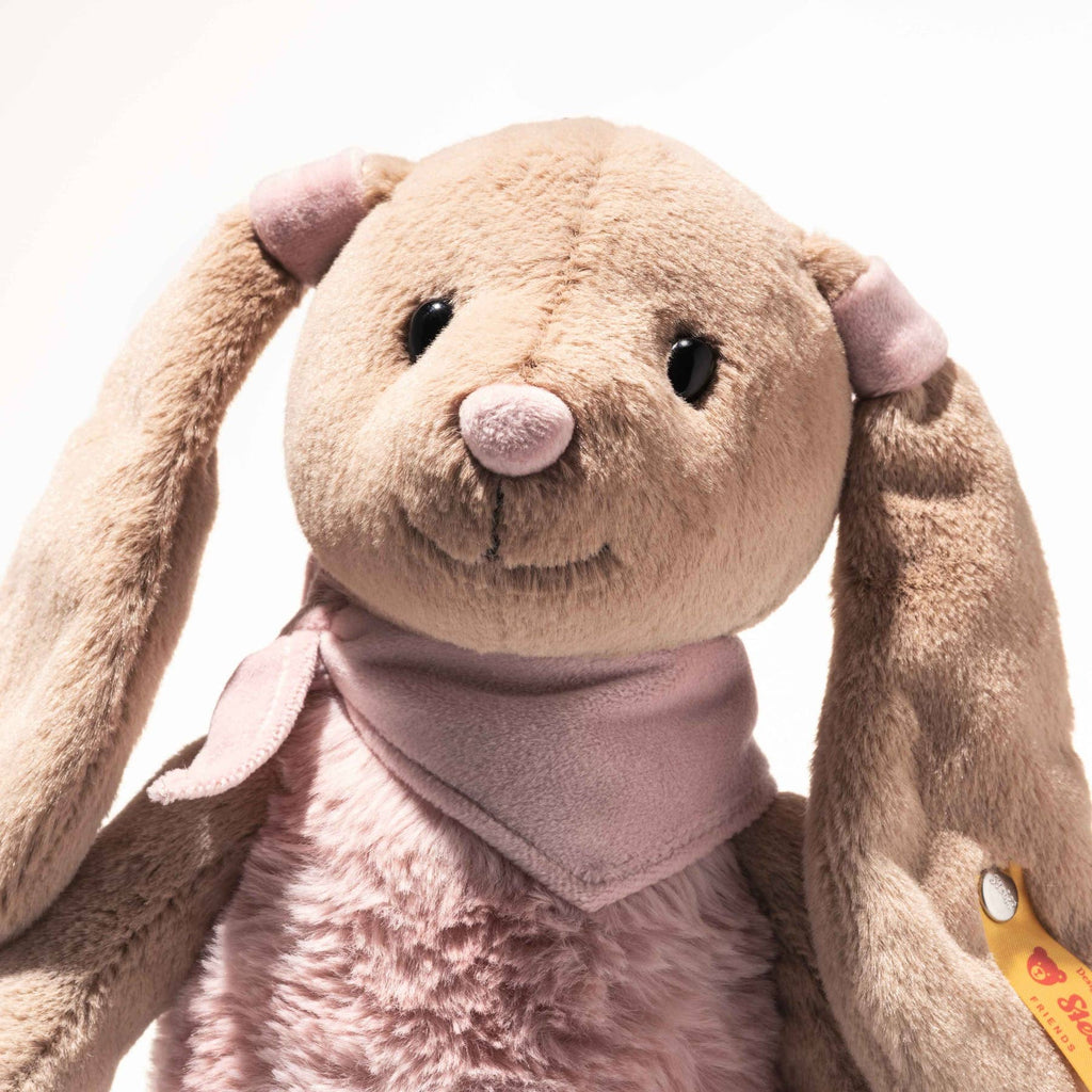 The Steiff Cowgirl Hoppie Rabbit is a soft, plush bunny with light brown fur, long floppy ears, black button eyes, a pink nose, a light pink scarf around its neck, and a yellow tag on its left ear.