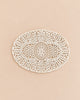 An oval-shaped, intricately patterned white lace doily, reminiscent of the Maileg Cozy Corner Set, is displayed on a smooth, light beige surface.