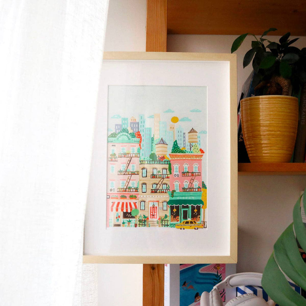 The Paint By Number Kit - New York framed illustration features vibrant city buildings with shops and apartments, displayed on a wooden shelf next to a potted plant and partially hidden by a white curtain.