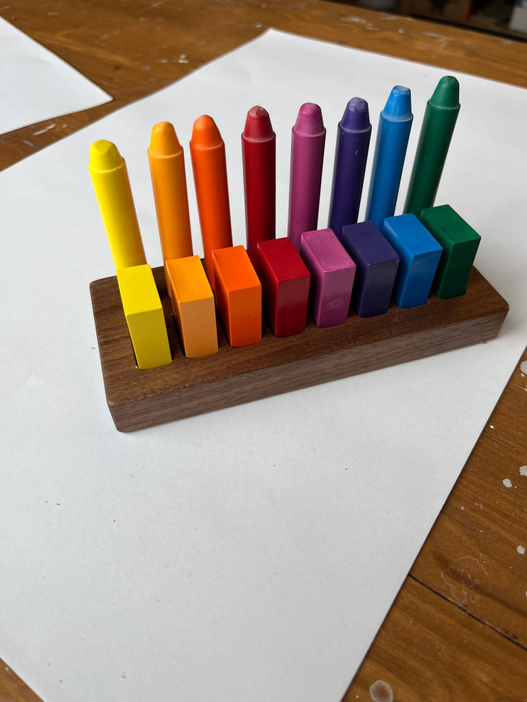 Colorful Filana Stockmar crayons arranged in a Wooden Crayon Holder 8 Stick / 8 Block on a table, representing a vibrant spectrum from yellow to blue colors. The background features a blank sheet of paper.