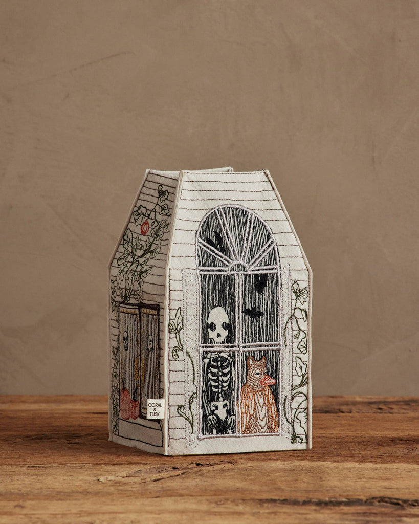 The Coral & Tusk Haunted House Tissue Box Cover is shaped like a house with embroidered skeleton, owl, and cat in the windows, climbing vines on the sides, and sits on wood with a textured brown background.