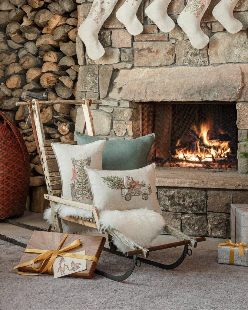 A cozy living room with a lit fireplace, stacked firewood, Christmas stockings on a stone mantel, woodland animal holiday decor, a Coral & Tusk Christmas Tree Car Pocket Pillow on a rustic sled, gold-bowed gift, and furry white rug.