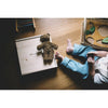 A small child sits on a wooden floor with toys nearby. A GOTS-certified Senger Naturwelt Stuffed Animal - Brown Bear rests on a light wooden box beside the child, who wears eco-friendly quality blue pants.
